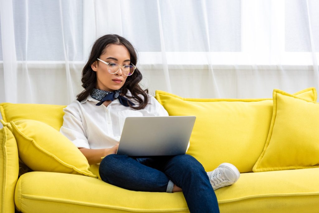 Serious asian freelancer in glasses using laptop while sitting comfortably on yellow sofa at home
